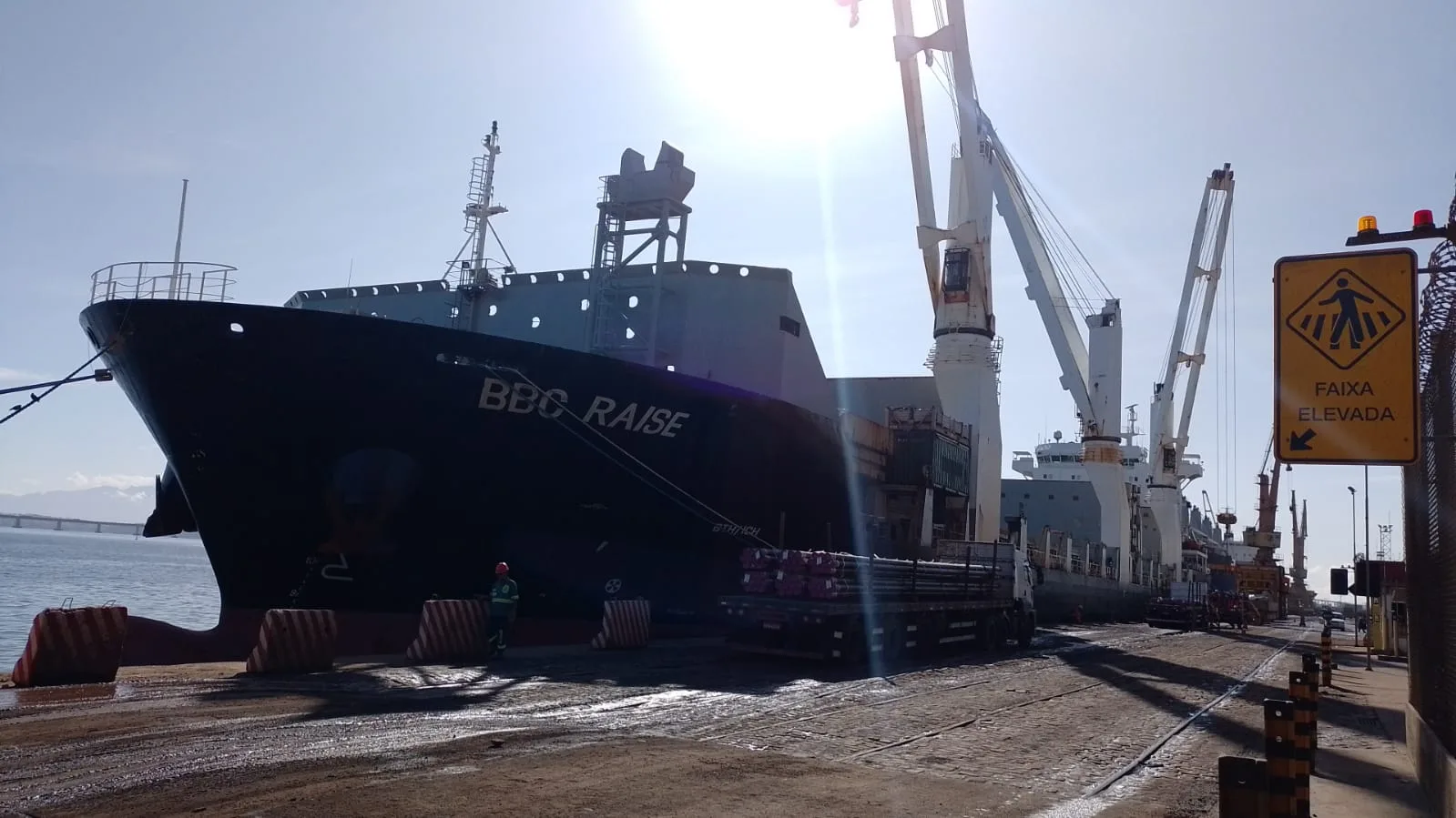 Navio break bulk BBC Raise atracado em terminal portuário brasileiro, com guindastes de bordo carregando carga de aço de um caminhão
