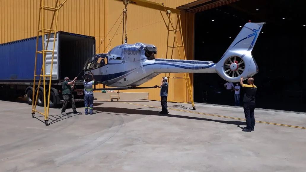 Helicóptero suspenso em pórtico de içamento sendo embarcado em contêiner dentro de um hangar