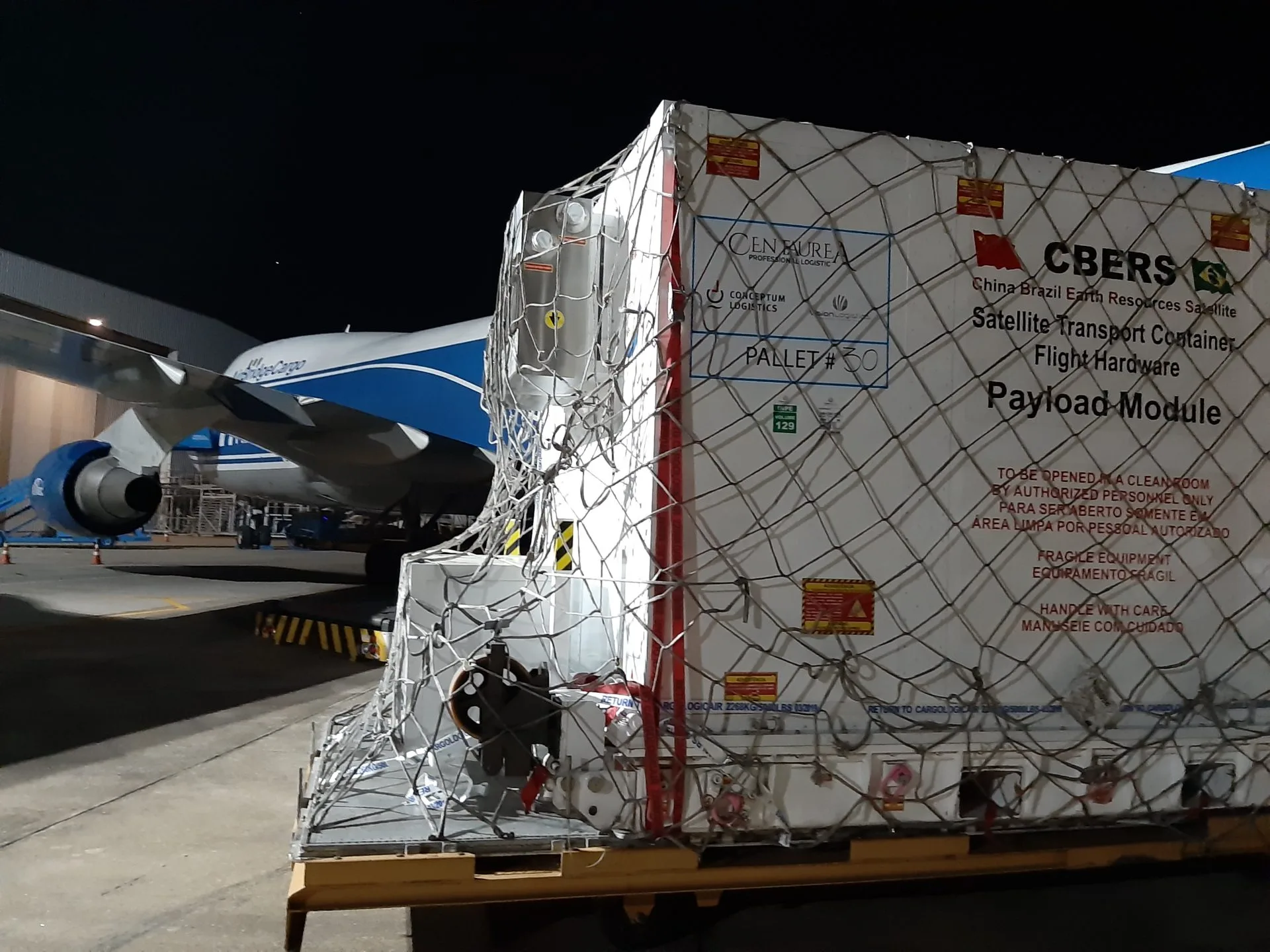 Contêiner de transporte de satélite sobre pallet ao lado de aeronave cargueira, à noite, em operação de fretamento aéreo